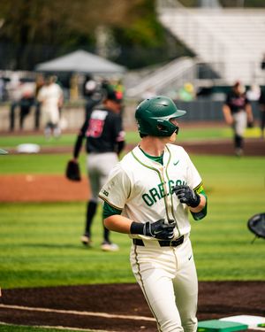 Oregon Ducks baseball player in white uniform and green helmet walks during a college baseball game.