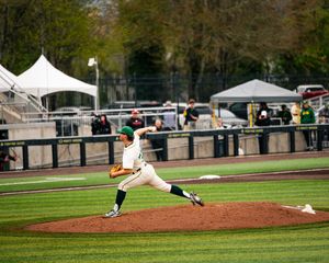Baseball pitcher in green and white uniform mid-throw on the mound during a daytime game at an outdoor stadium.