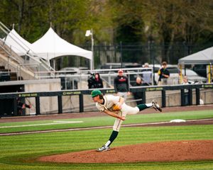 Baseball pitcher in green and white uniform delivers a pitch on an outdoor college baseball field.