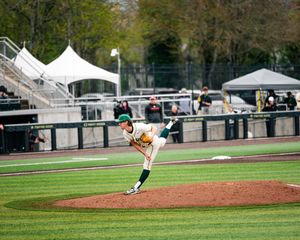 Baseball pitcher in green and yellow uniform mid-throw on a manicured mound during a collegiate game.