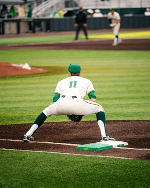 Baseball player #11 in white and green uniform stretches at first base during a college baseball game on a green field.