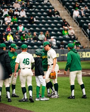 Oregon Ducks baseball team huddles on the field during a game at a stadium with fans in the stands.