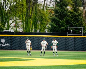 Three baseball players stand in the outfield during a game at a green stadium with tree-lined backdrop.