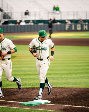 Oregon Ducks baseball players in white uniforms running bases during a college baseball game at a green stadium.