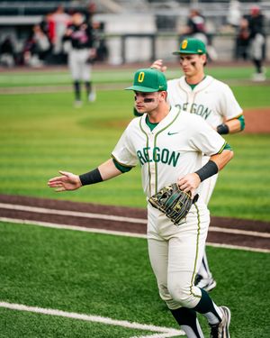 Oregon Ducks baseball player in white uniform with green accents walks on field holding glove during college game.