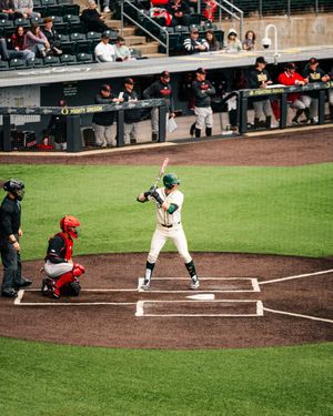 Baseball batter in green uniform stands at home plate with catcher and umpire during a college baseball game.