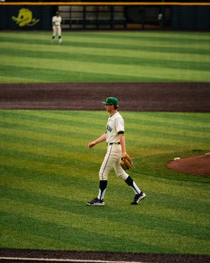 Baseball player in green and white uniform walks on field holding glove, with teammate visible in outfield background.