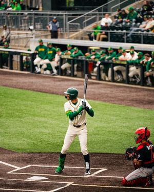 Baseball batter in green uniform stands ready at plate with catcher and dugout visible at outdoor stadium.