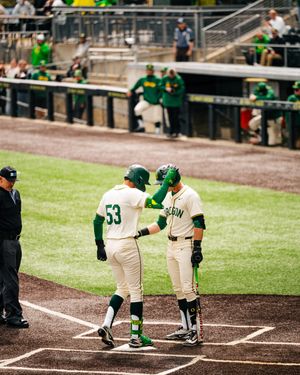 Two baseball players in green and white uniforms celebrate at home plate during a college baseball game.