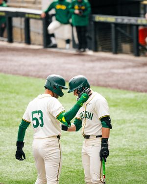 Two Oregon Ducks baseball players in green helmets and white uniforms celebrate near home plate during a college game.