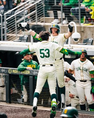 Oregon Ducks baseball players in green uniforms celebrate on field, player #53 jumps with teammate after scoring a run.