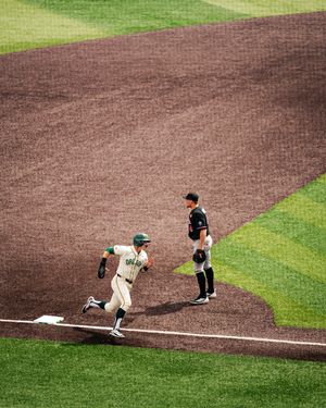 Baseball player in green uniform rounds third base as fielder stands nearby on a dirt infield during a game.