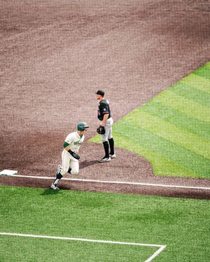 Baseball player crouching at first base in green uniform while opponent in black stands nearby on dirt infield.