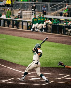 A baseball batter in white and green uniform swings at home plate during a college game with dugout visible in background.