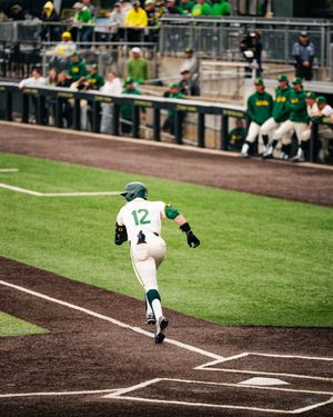 Baseball player #12 in green and white uniform rounds the bases during a college baseball game.