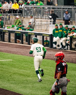 Baseball player #53 in green uniform runs bases while red-uniformed catcher stands nearby during college baseball game.