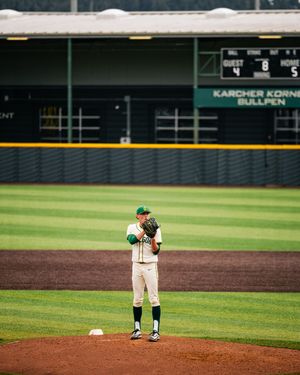 Baseball pitcher in white and green uniform stands on mound at Karcher Korner Bullpen stadium, scoreboard shows 4-5.