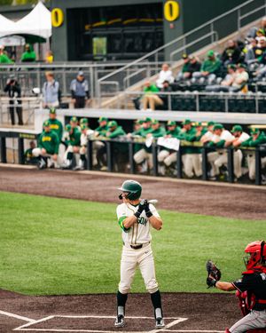 Oregon Ducks baseball batter stands ready at home plate in green uniform during a college baseball game at a packed stadium.