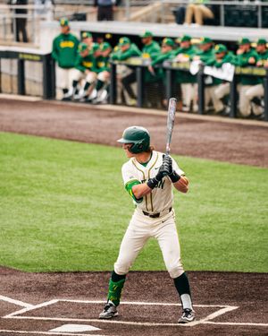 Baseball player in green uniform batting at home plate with teammates in dugout behind him.