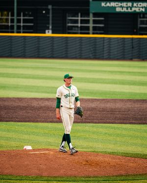 Oregon Ducks baseball pitcher stands on the mound in white and green uniform at a college baseball stadium.