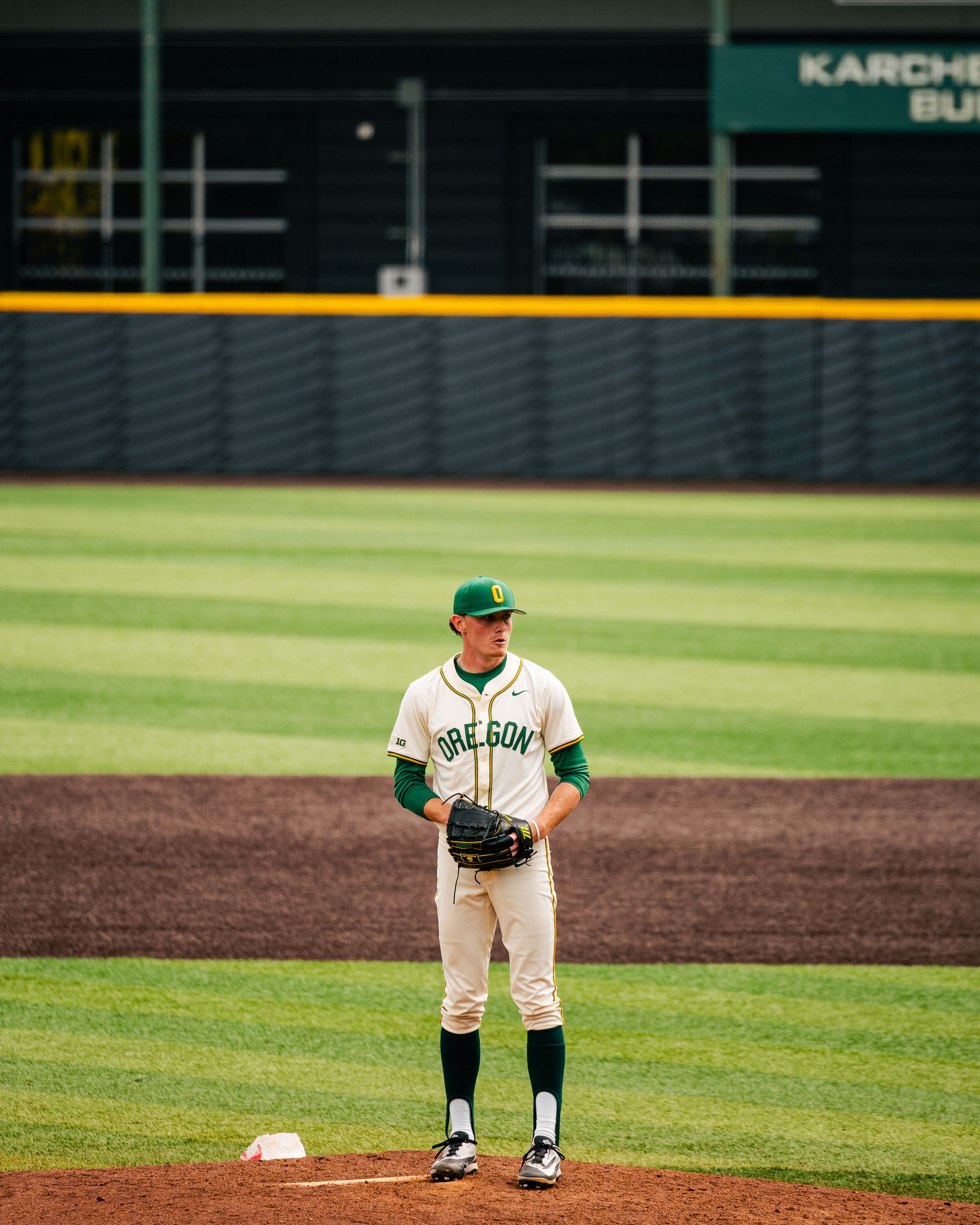 Oregon Ducks baseball pitcher stands on the mound in white and green uniform at PK Park stadium.