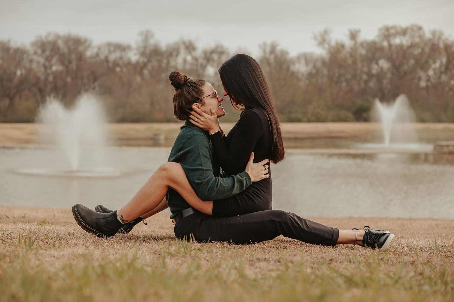 Two women sitting on grass by a pond with fountains, sharing a romantic embrace and kiss outdoors.