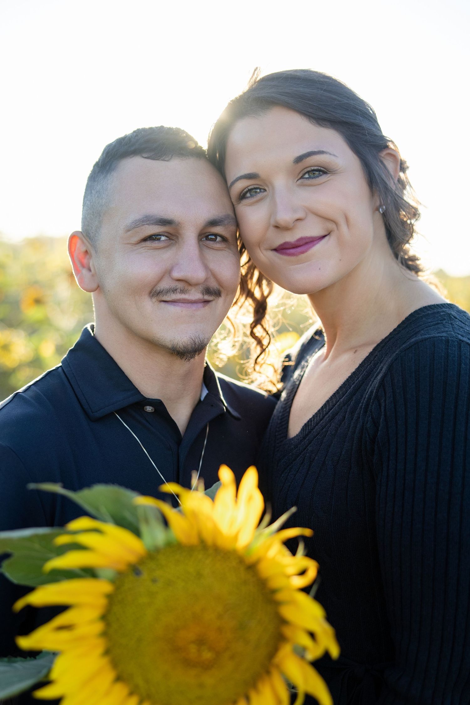 A smiling couple poses together in a sunflower field outside outside of pensacola, florida. Both man and woman are wearing black shirts