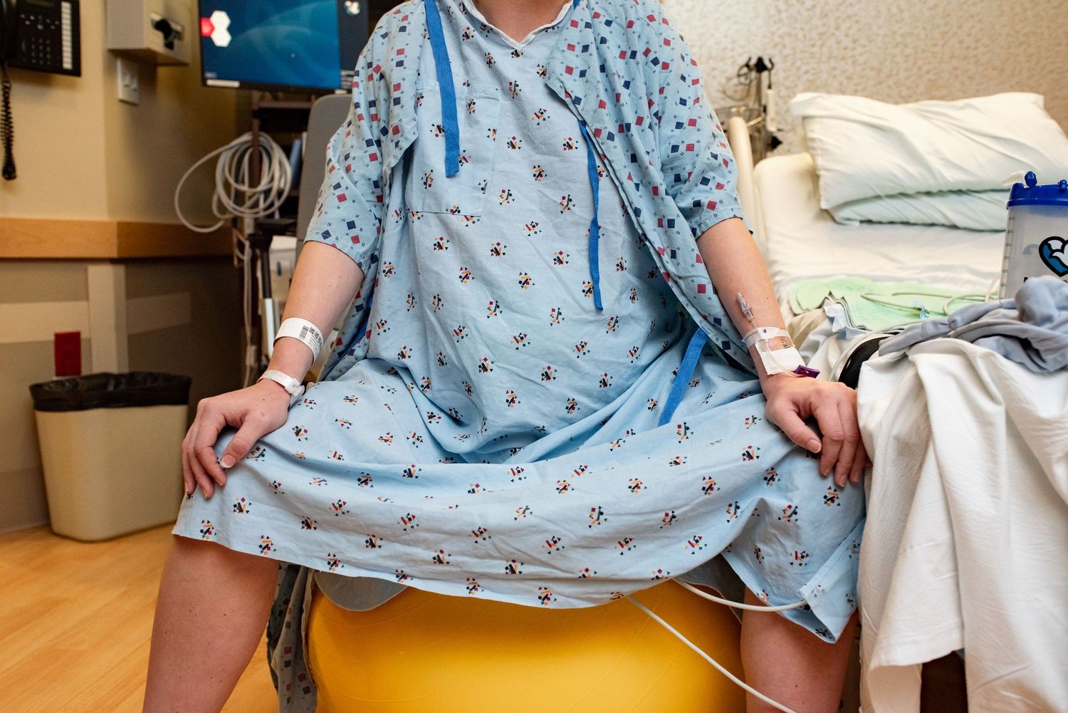 Hospital patient in gown sitting on yellow exercise ball during recovery in medical room.