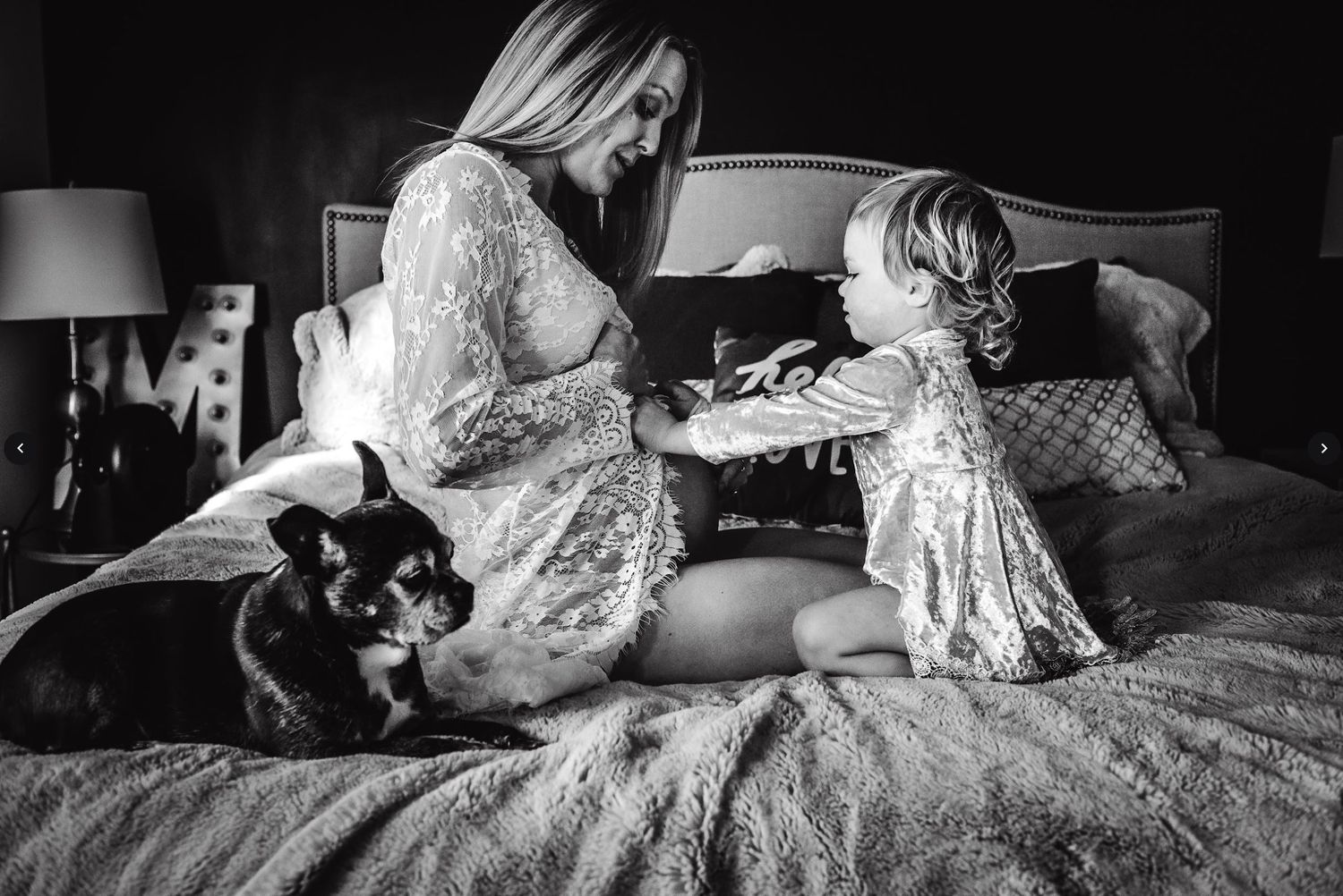 Black and white maternity image of a tender moment on a bed with a small dog and toddler