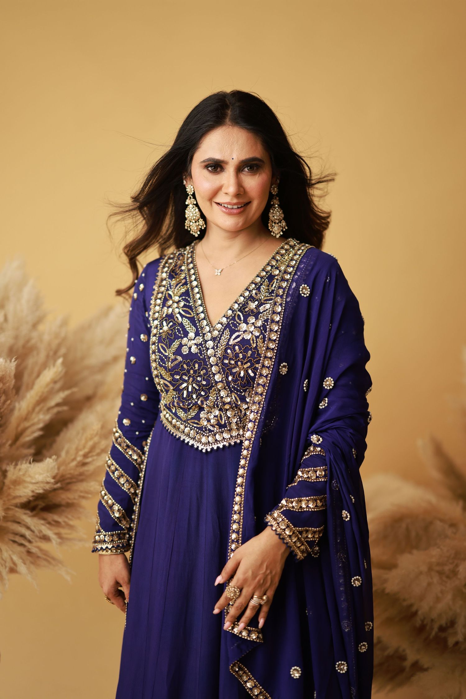 A deep blue embroidered Indian anarkali dress with intricate gold embellishments and matching jewelry against a beige backdrop.