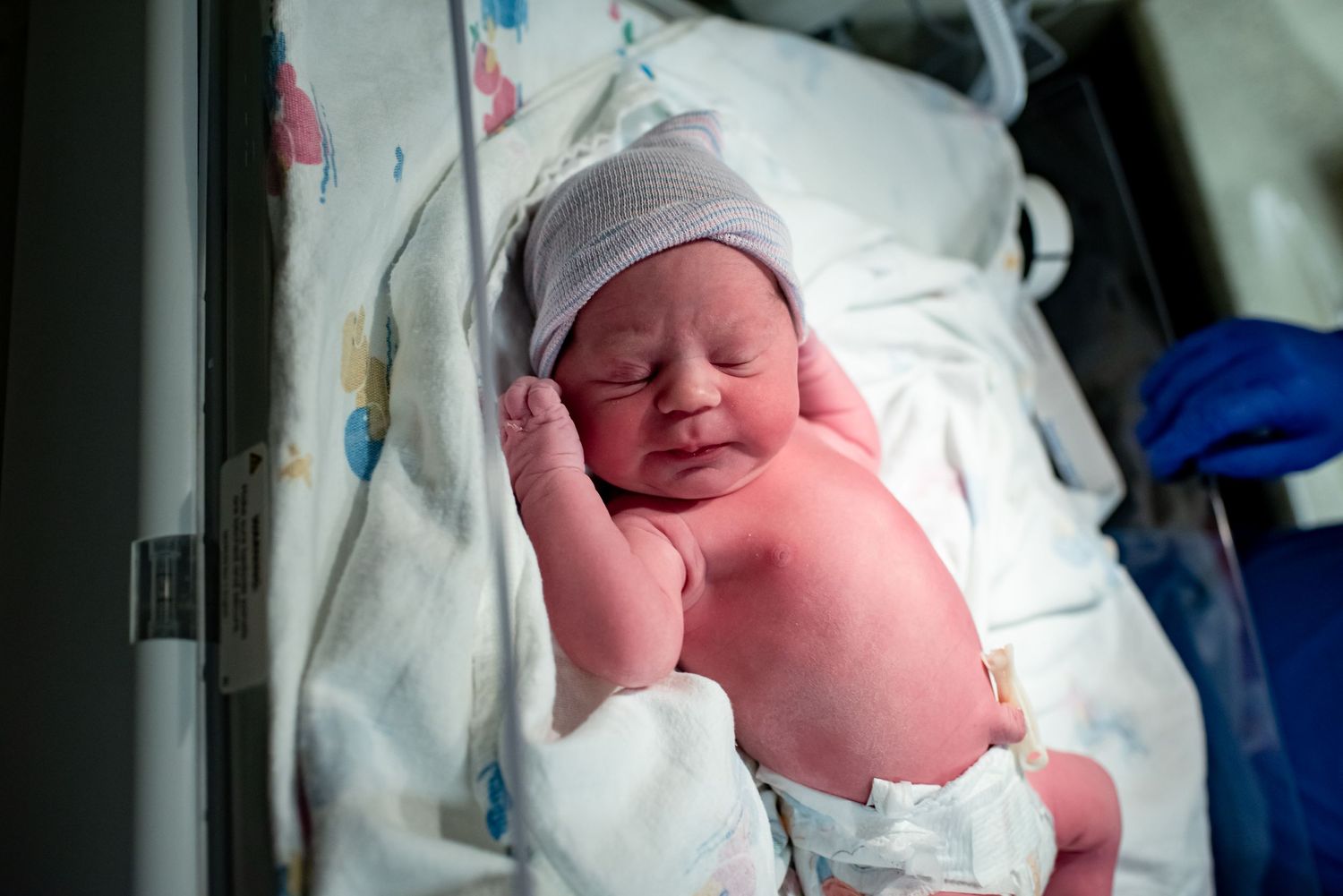 Newborn baby wearing a knit cap rests peacefully on white hospital bedding.