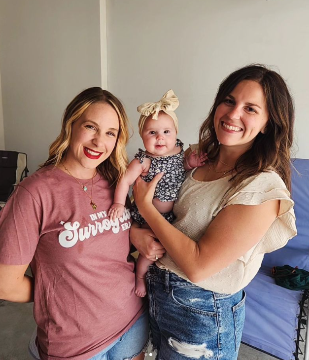 Three people pose together indoors, with a small baby wearing a floral headband in the center.