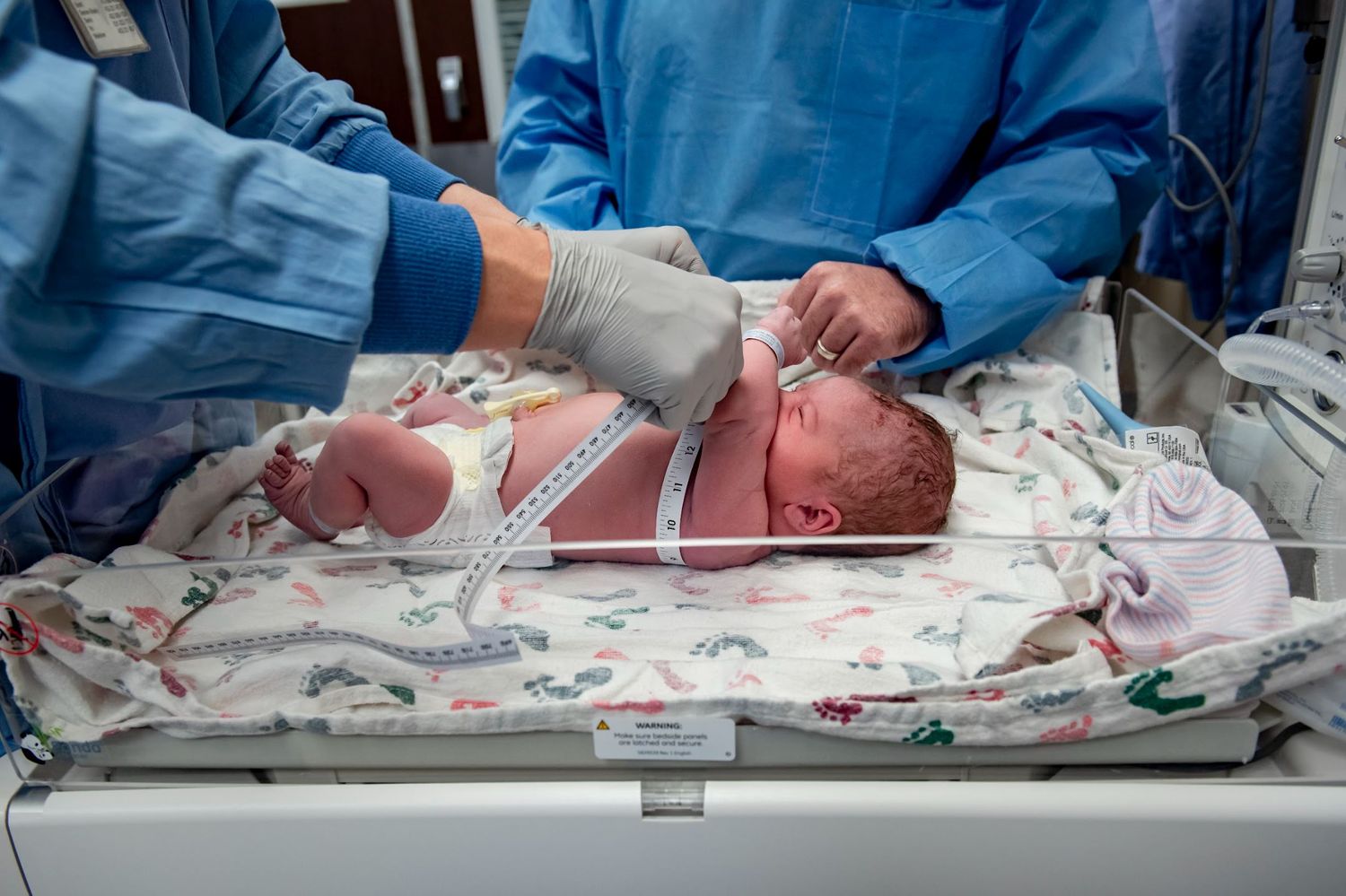 Medical staff examining newborn baby on hospital examination table with monitoring equipment.
