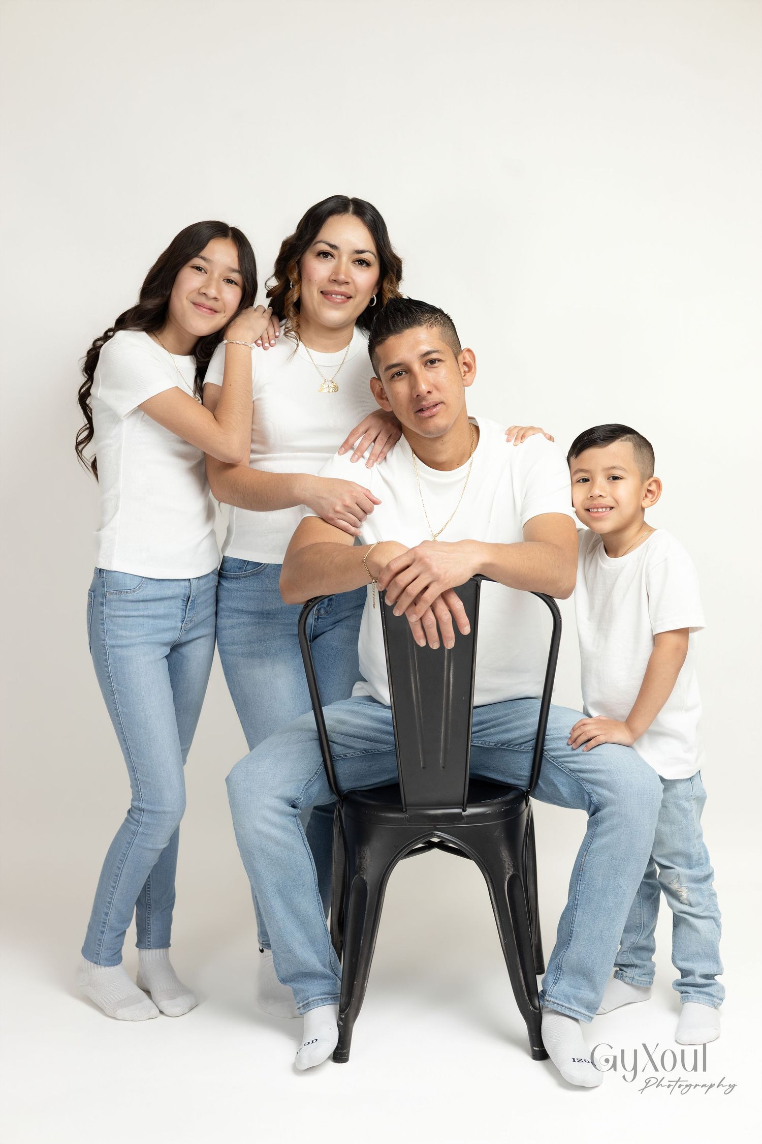 Family of four in matching white shirts and jeans posing together in a professional studio portrait photo.