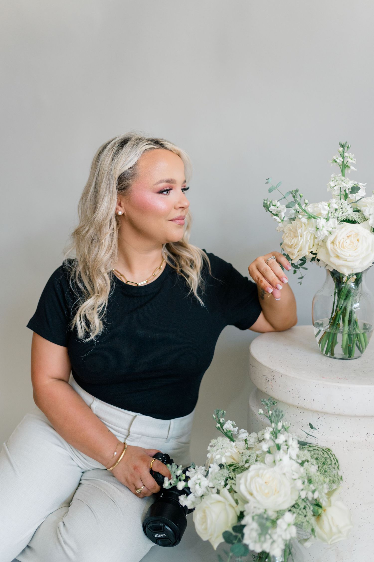 Blonde woman in black tee holds camera beside white roses in glass vase on marble table.