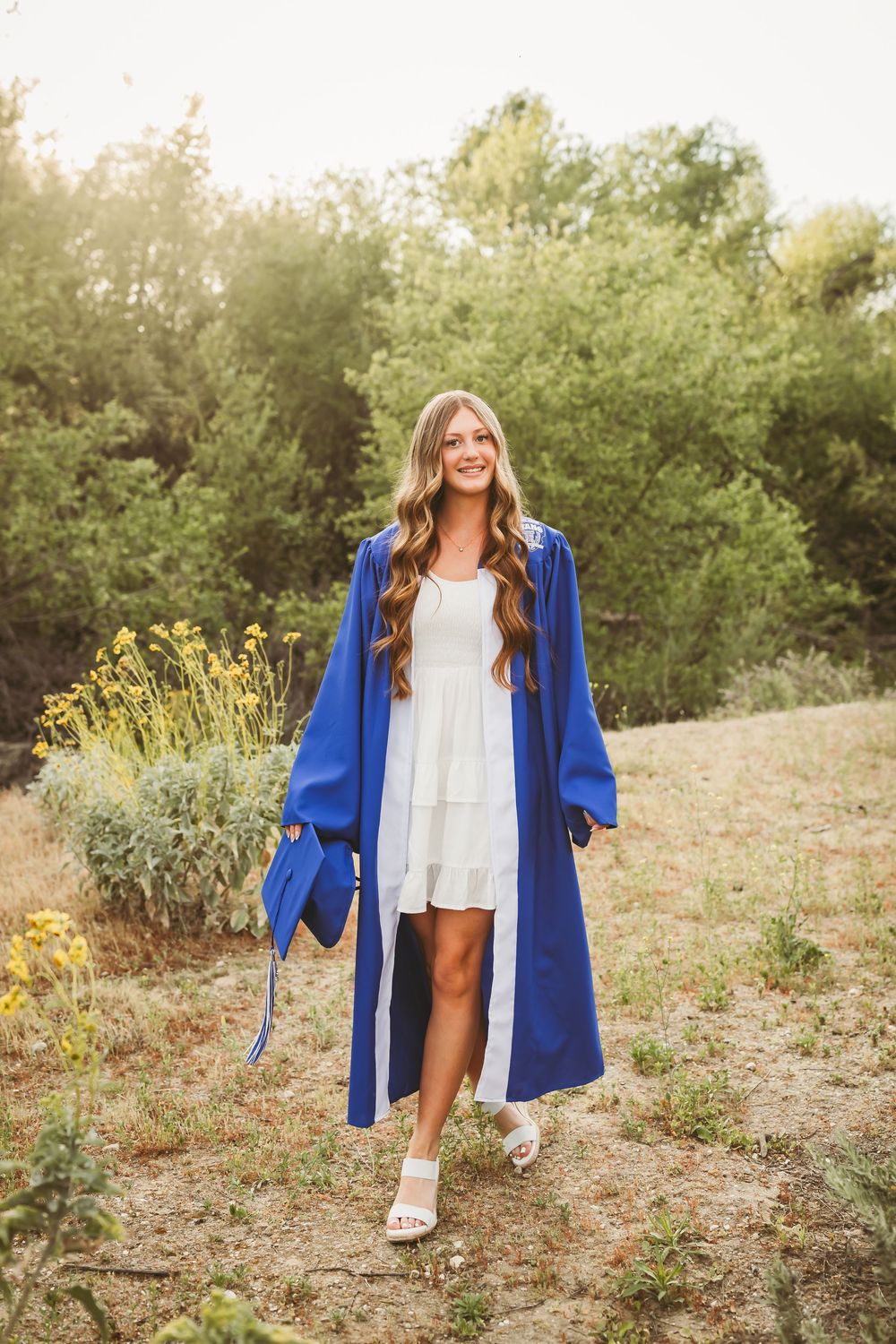 Young woman in blue graduation gown holding cap, wearing white dress, smiling outdoors among green trees and yellow flowers.