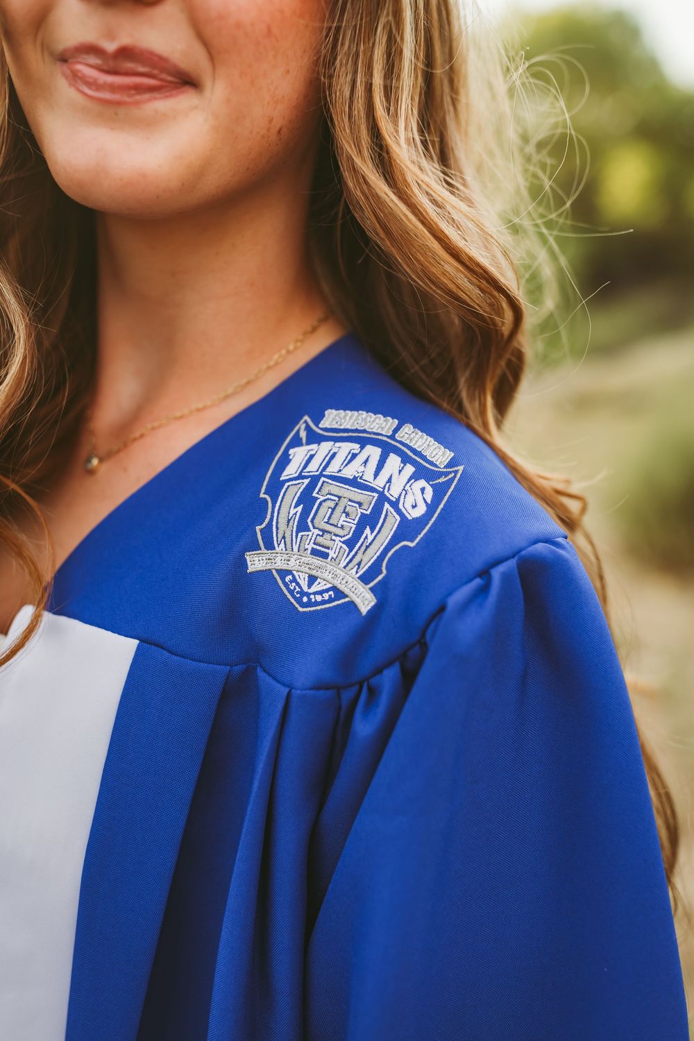 Young woman in royal blue Titans graduation gown with embroidered crest, smiling outdoors.