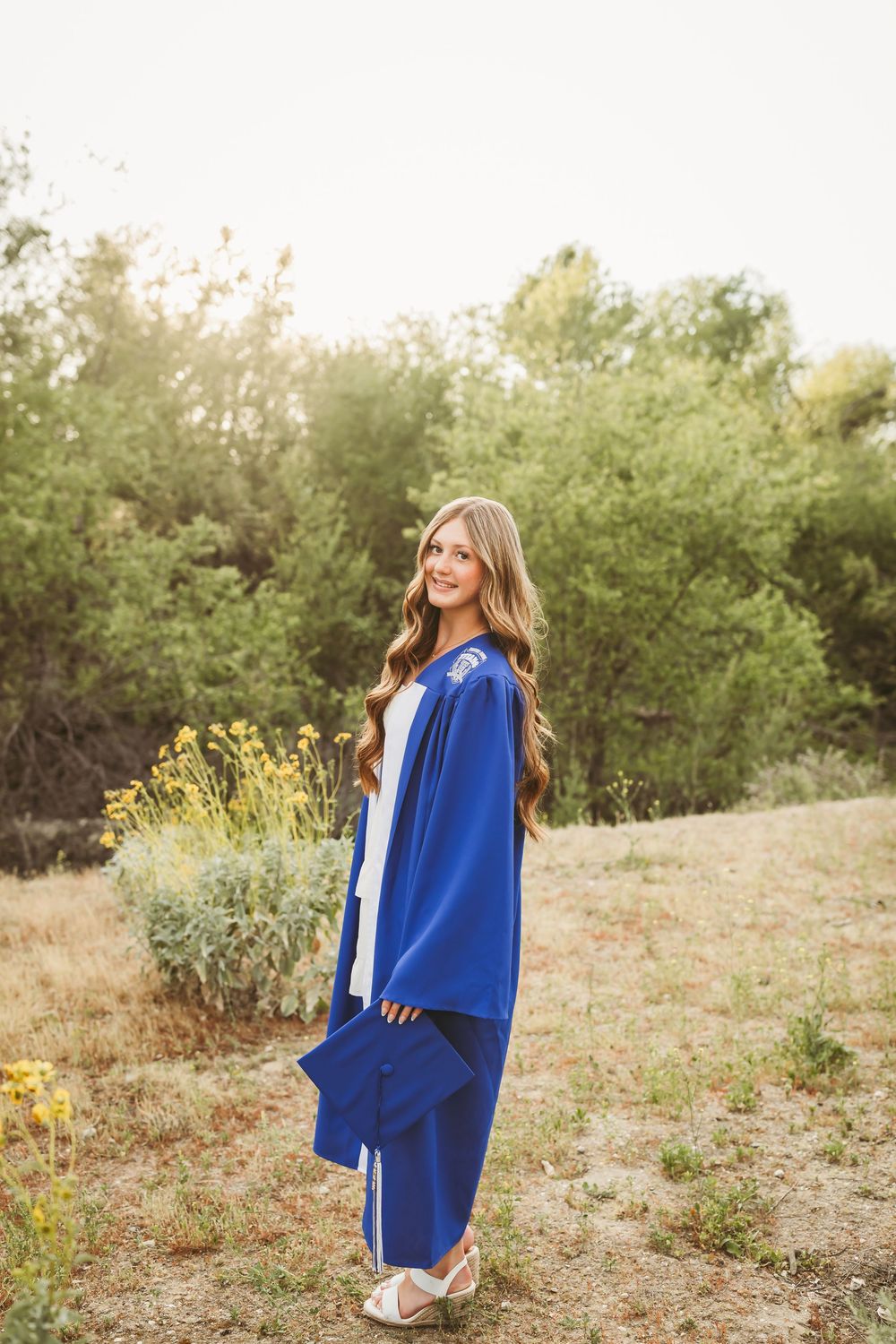 Young woman in blue graduation cap and gown smiling outdoors in a natural setting with wildflowers and trees.