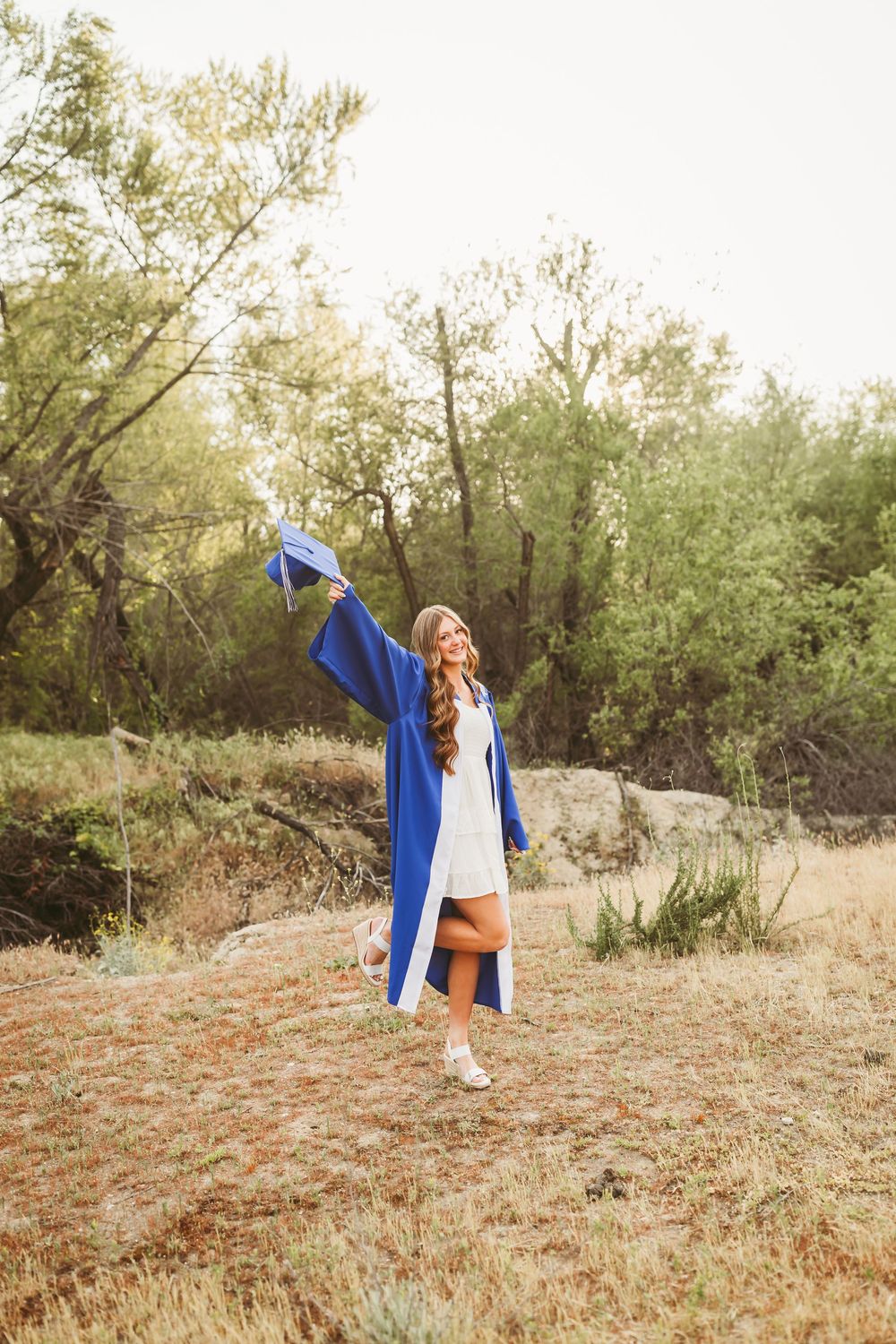 Young woman in blue graduation gown raises cap triumphantly outdoors in a field surrounded by green trees.