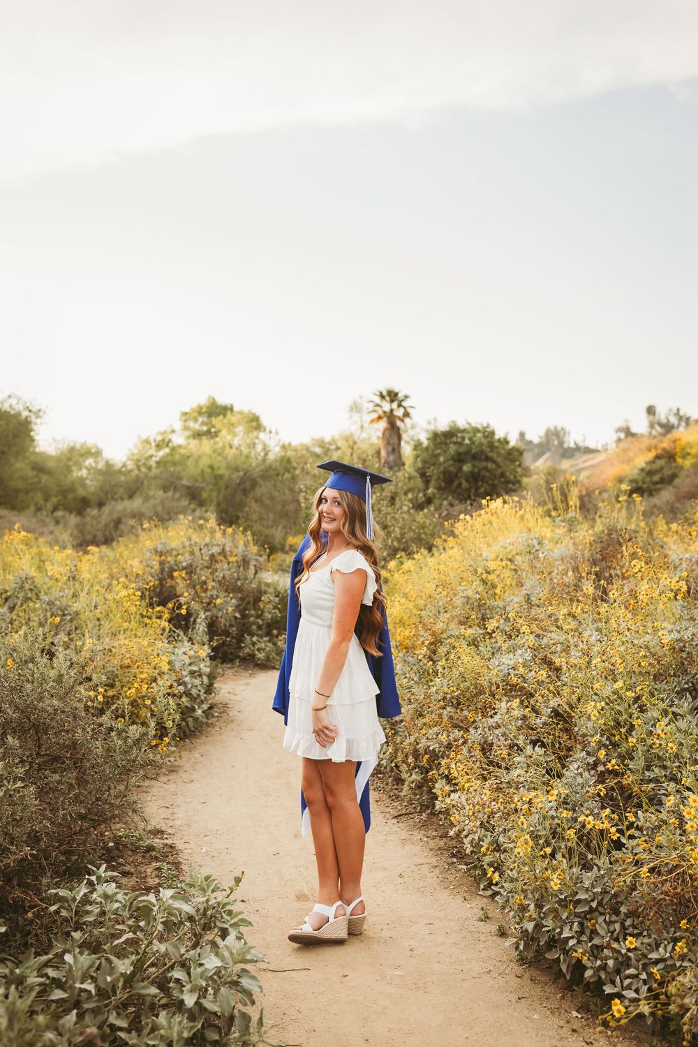 A smiling graduate in blue cap and gown poses on a nature trail surrounded by yellow wildflowers.