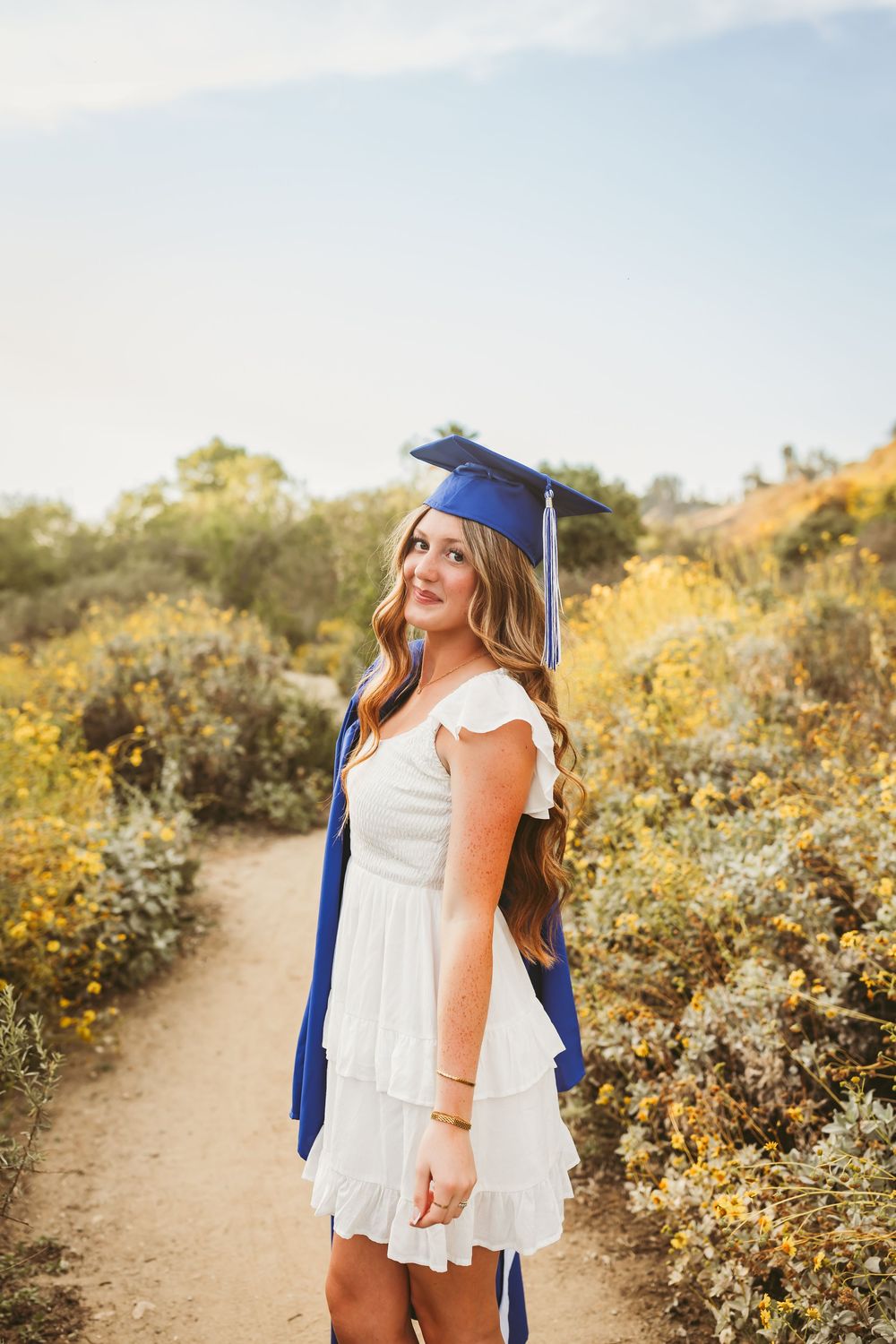 Young woman in white dress and blue graduation cap poses on a scenic trail surrounded by wildflowers.
