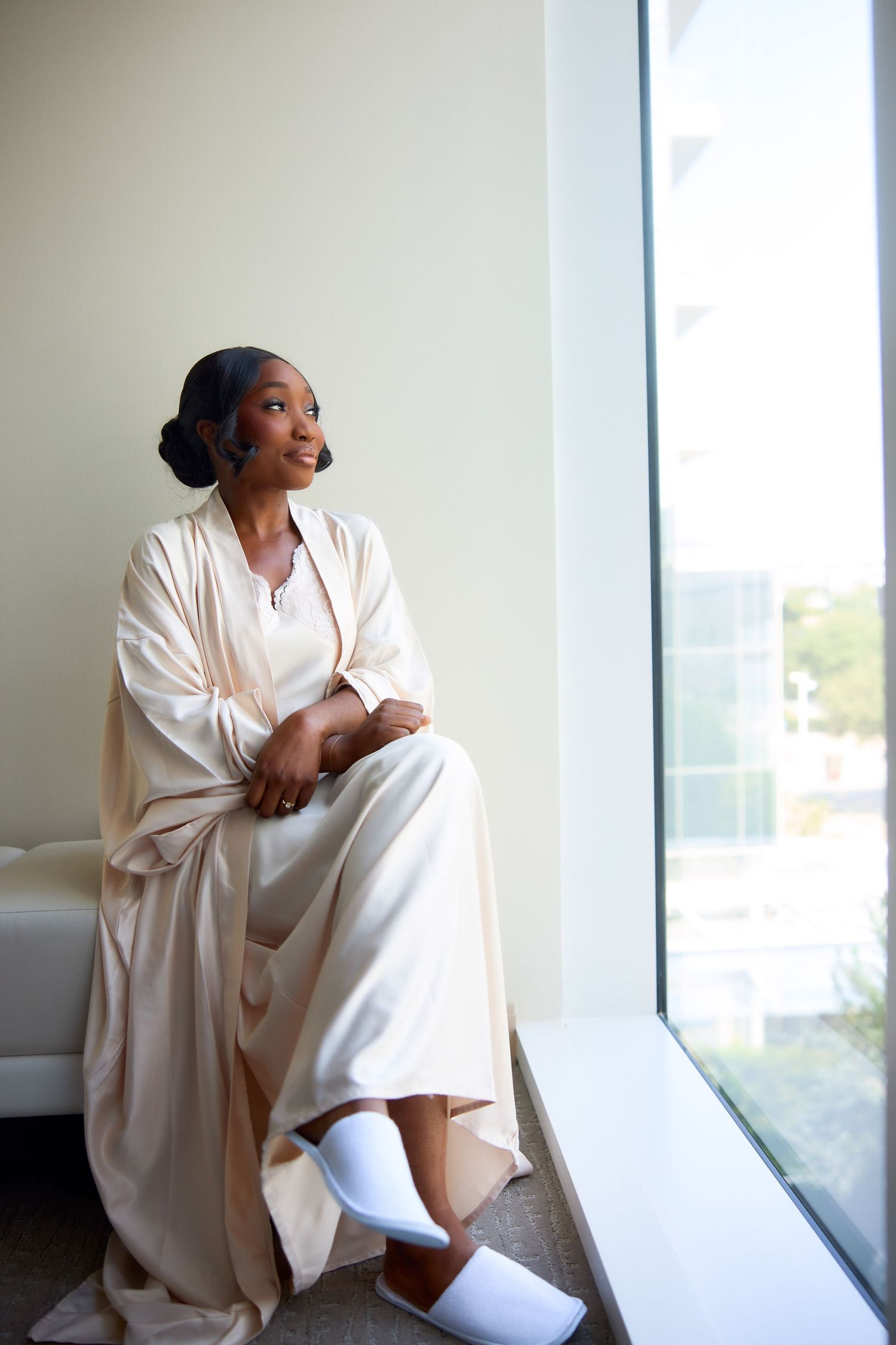 A young Black woman in a cream satin robe and white slippers sits by a large window, smiling gracefully.