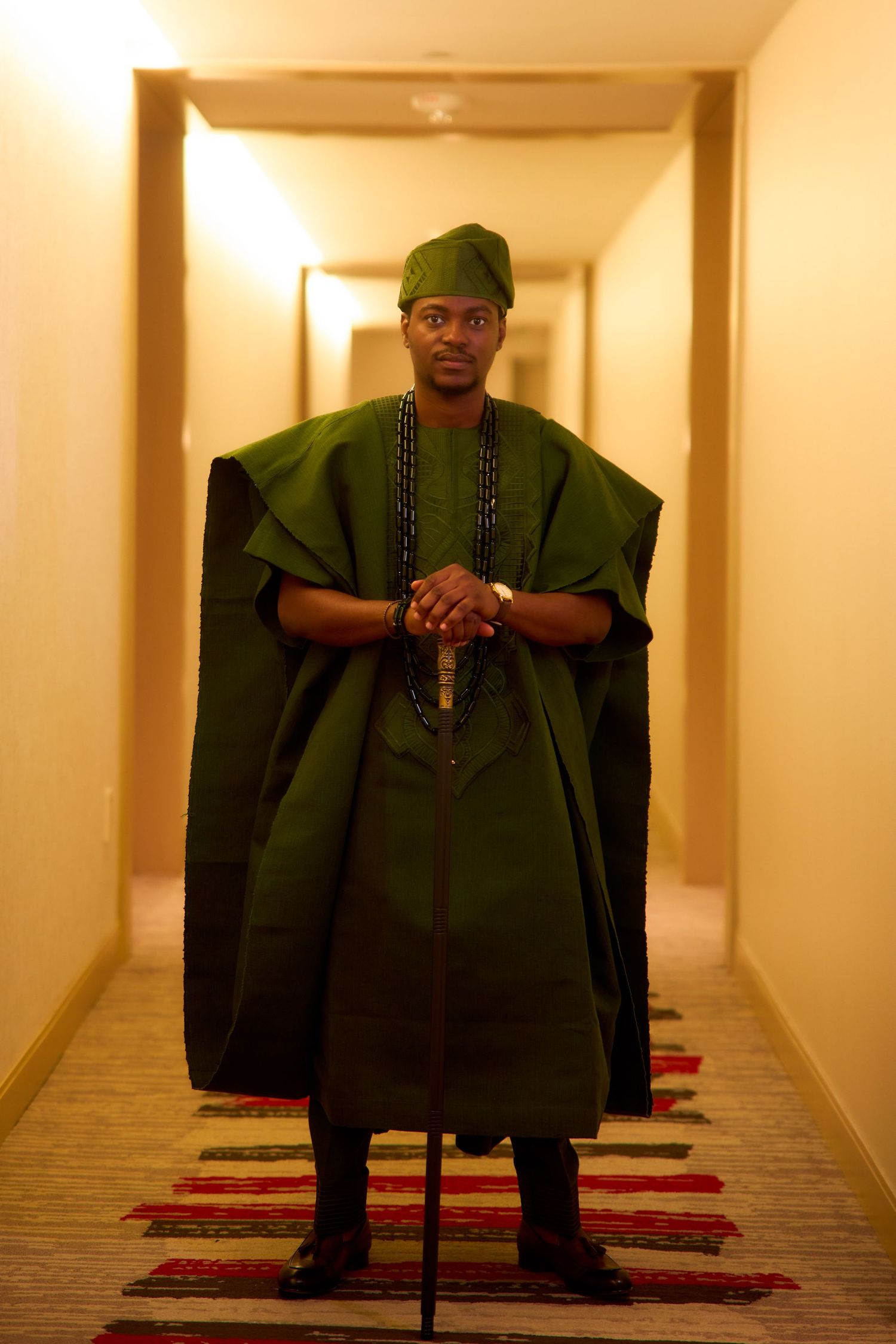 Man in traditional green African agbada attire with matching cap, holding a cane in a hotel corridor.