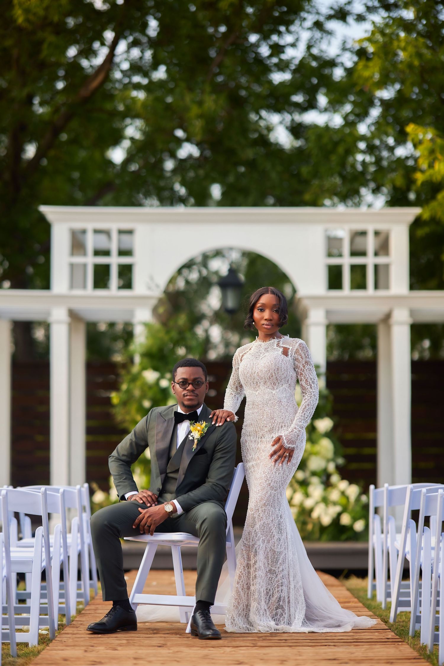 Elegant Black couple posing at outdoor wedding ceremony, groom in gray suit, bride in white lace gown, garden backdrop.