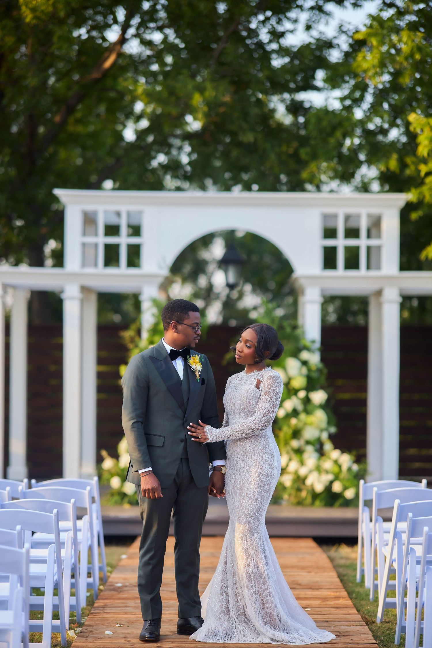 Elegant Black couple in wedding attire standing at outdoor ceremony aisle with white chairs and floral arch backdrop.