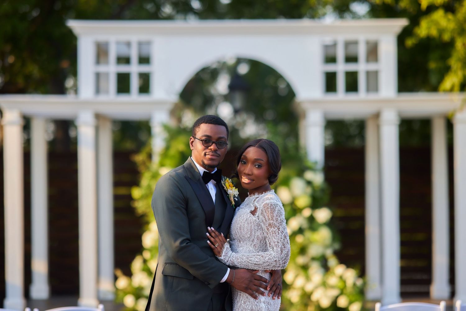 Elegant Black couple posing at outdoor wedding ceremony with white arch gazebo backdrop.