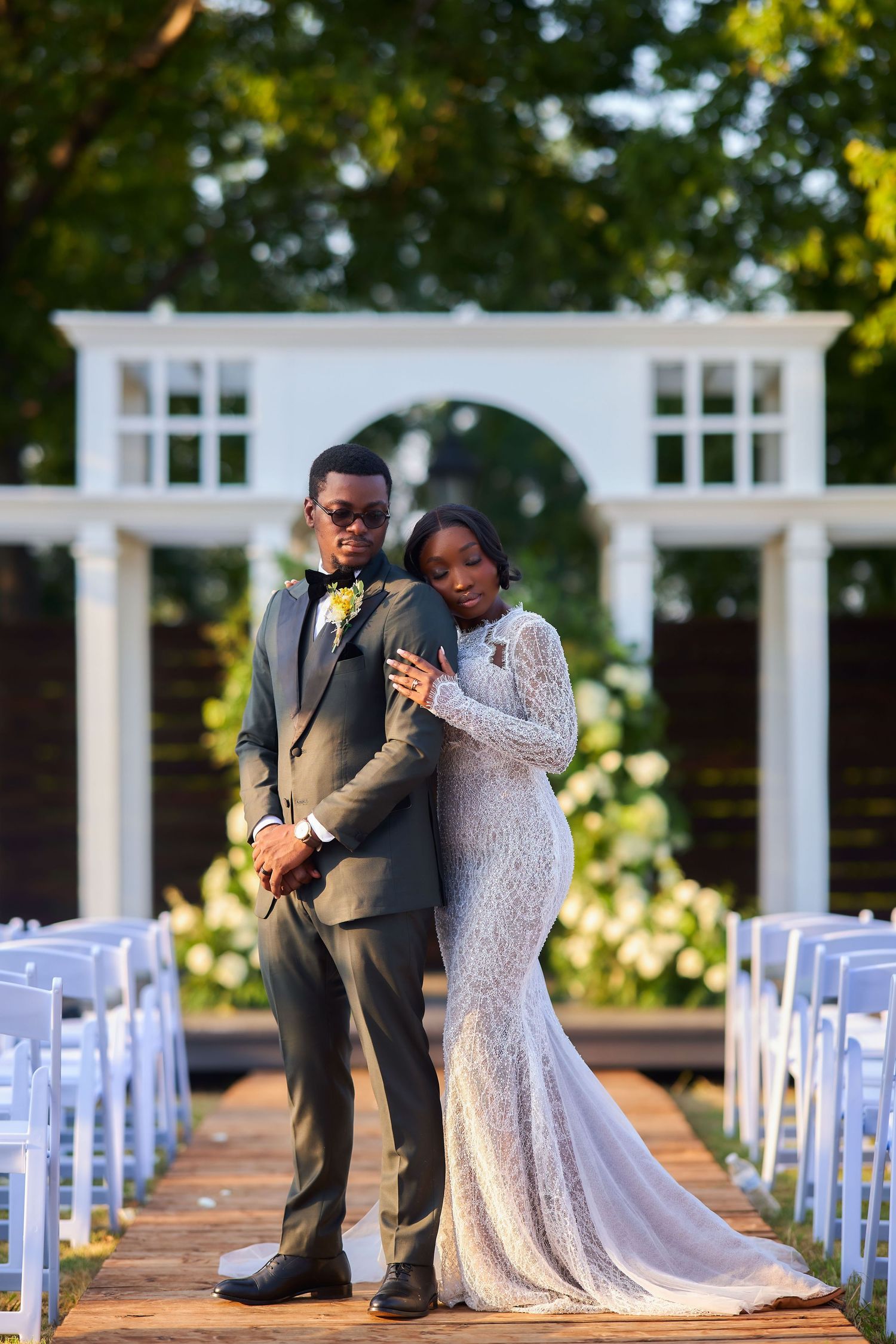 Elegant bride and groom posing at outdoor wedding ceremony aisle with white arch and chairs.