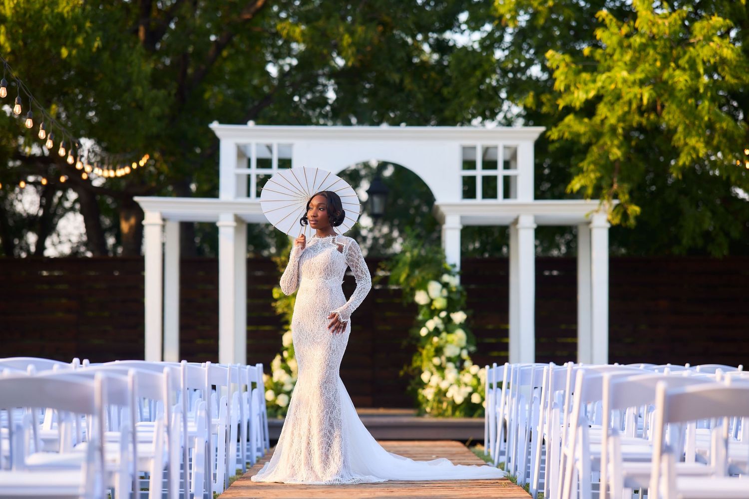 Bride in white lace gown poses at outdoor wedding venue with white chairs and floral arch backdrop.