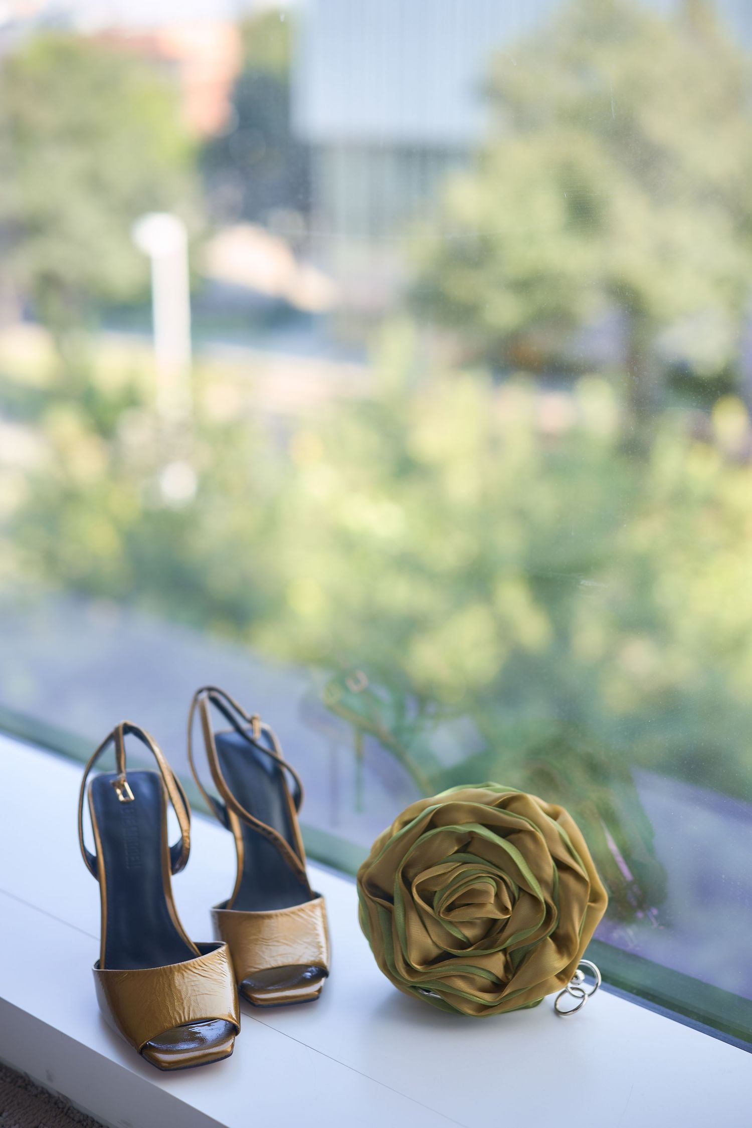 Gold strappy heels and green fabric rose corsage displayed on white windowsill with blurred garden backdrop.