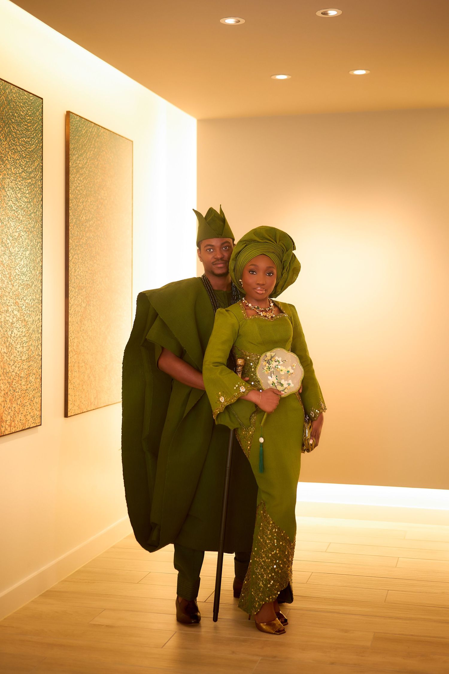 Couple in matching green traditional African attire posing elegantly in a warmly lit hallway.